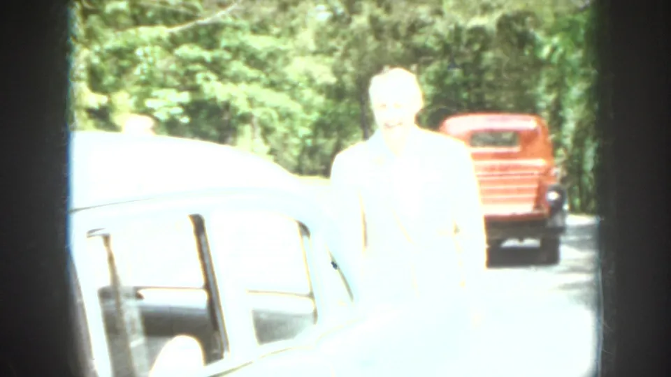 Washed-out 1958 Virginia home-movie frame showing a person beside a light-colored car with a red truck farther back.