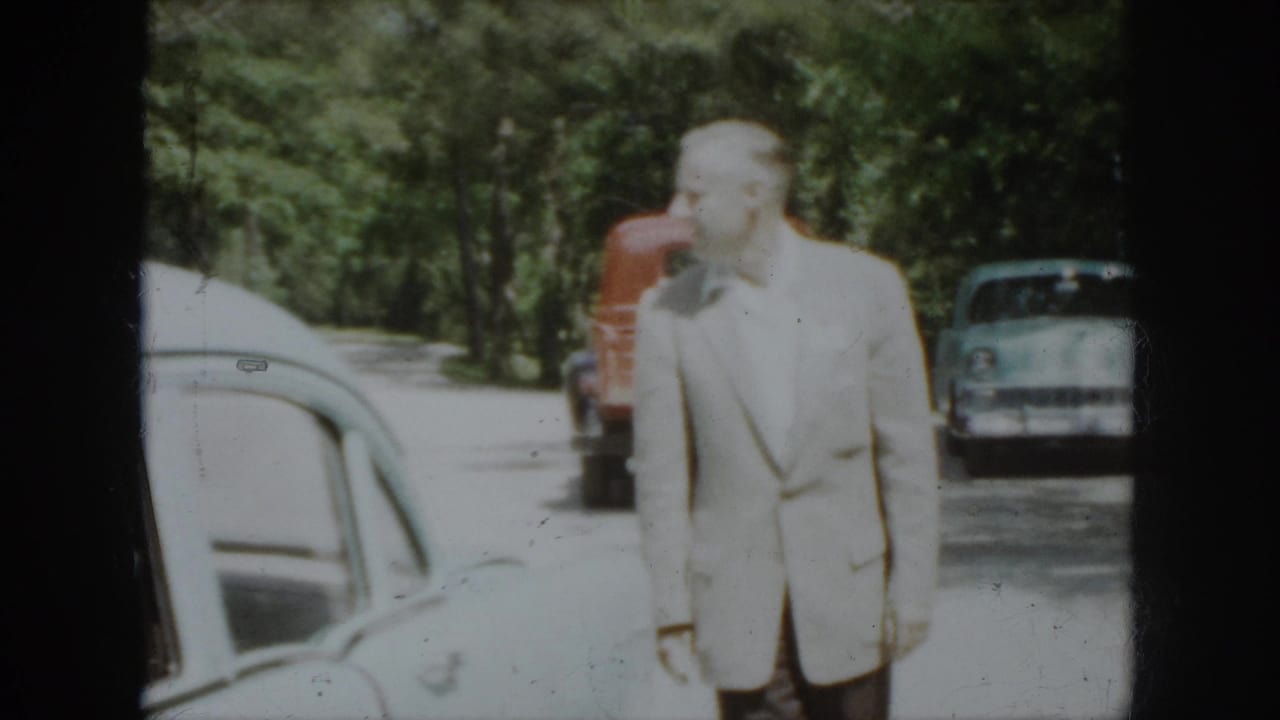 1958 Virginia frame showing a man in a light suit near parked mid-century cars and a red truck along a wooded edge.