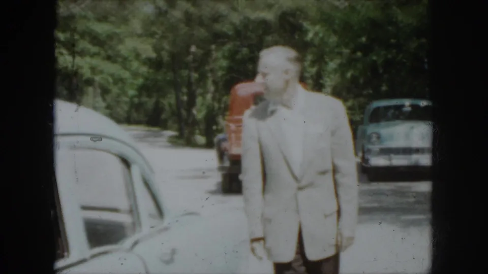 1958 Virginia frame showing a man in a light suit near parked mid-century cars and a red truck along a wooded edge.