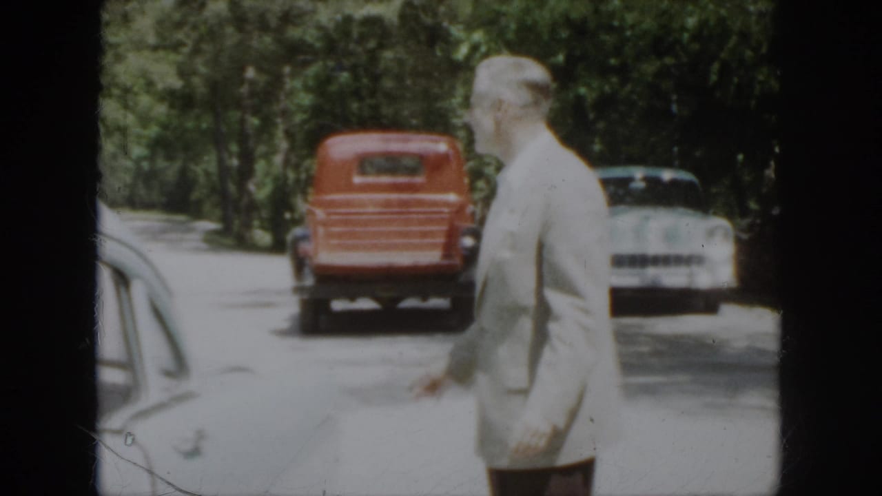 In 1958 Virginia home-movie footage, a suited man stands near parked mid-century vehicles on a shaded road.