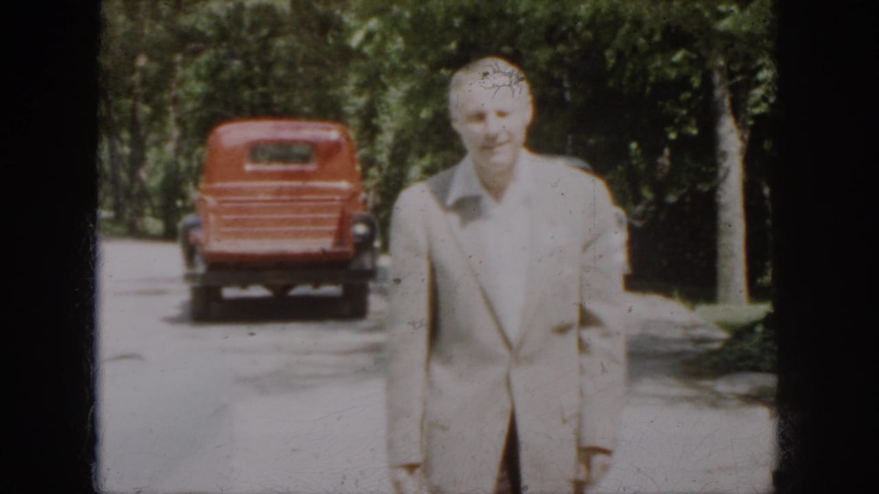 A man in a light-colored suit faces the camera on a Virginia roadside, with a red pickup and pale sedan behind him.