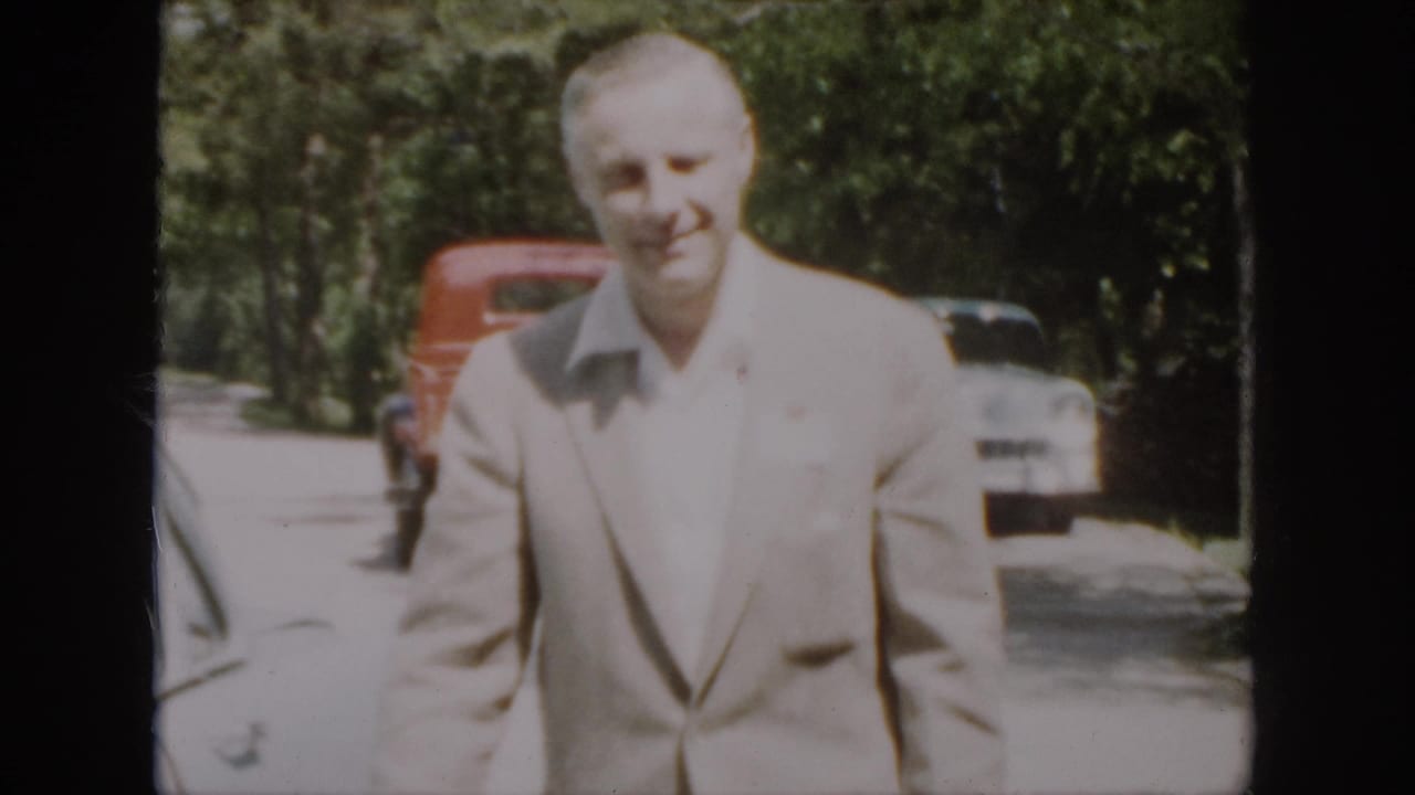 The suited man appears closer to the camera, smiling slightly as vintage cars remain parked along the wooded road.