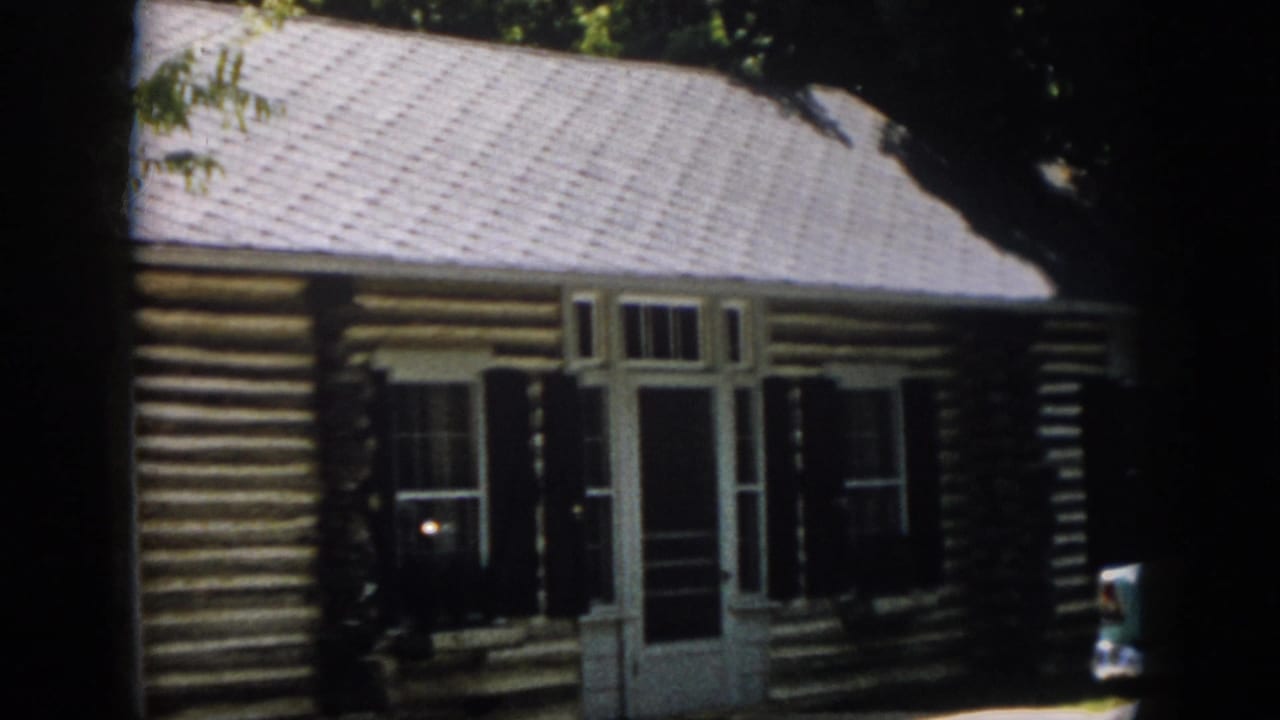 1958 Virginia 8mm frame showing a rustic log cabin front with white trim, screened doorway, dark shutters and leafy shade.