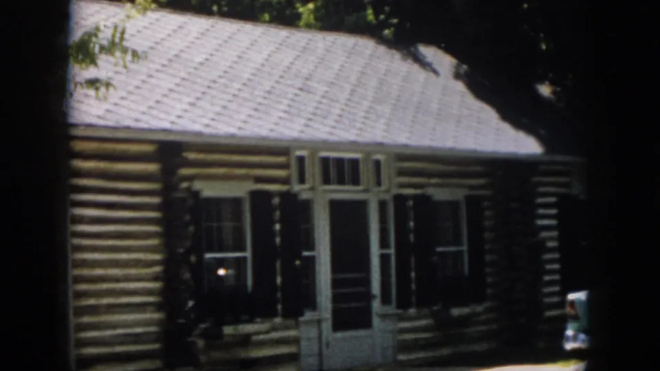 1958 Virginia 8mm frame showing a rustic log cabin front with white trim, screened doorway, dark shutters and leafy shade.