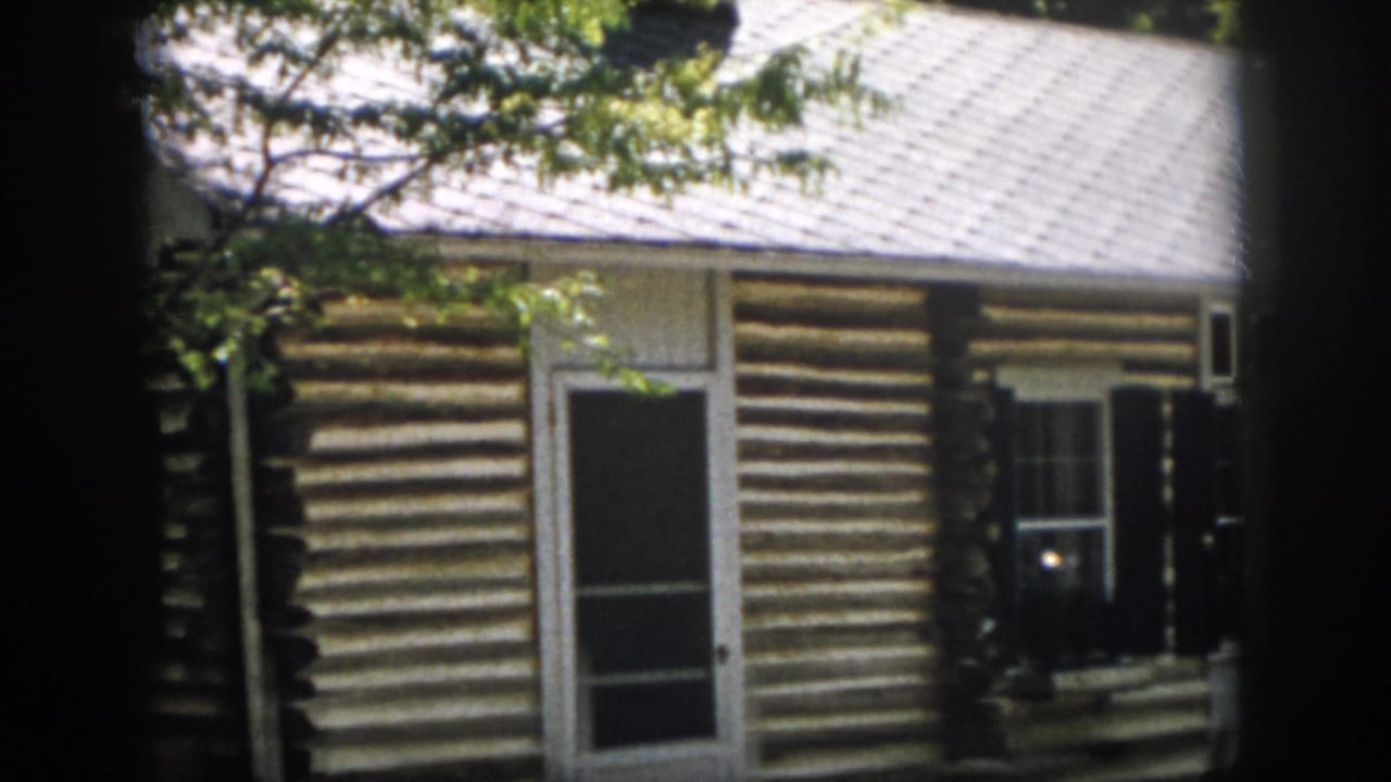 1958 Virginia home-movie frame of a log cabin exterior seen from a side-front angle, with screened entry and shuttered window.