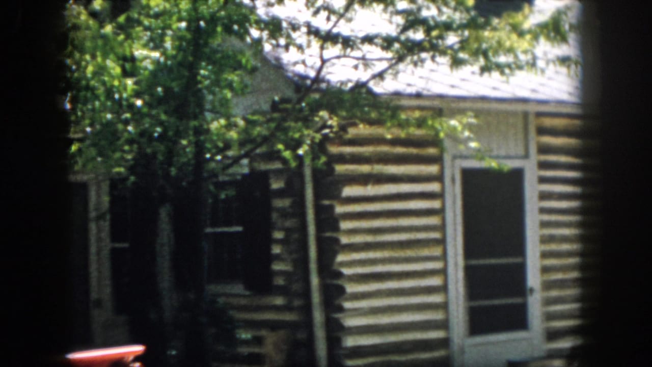 1958 Virginia 8mm frame with a tree partially blocking the rustic log cabin, white-trimmed door and shuttered window.