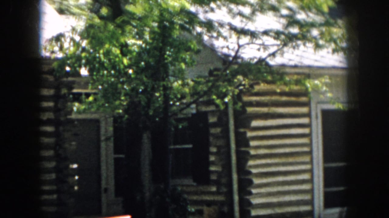 Rustic log cabin exterior with trees, windows, and dark film-frame edges.