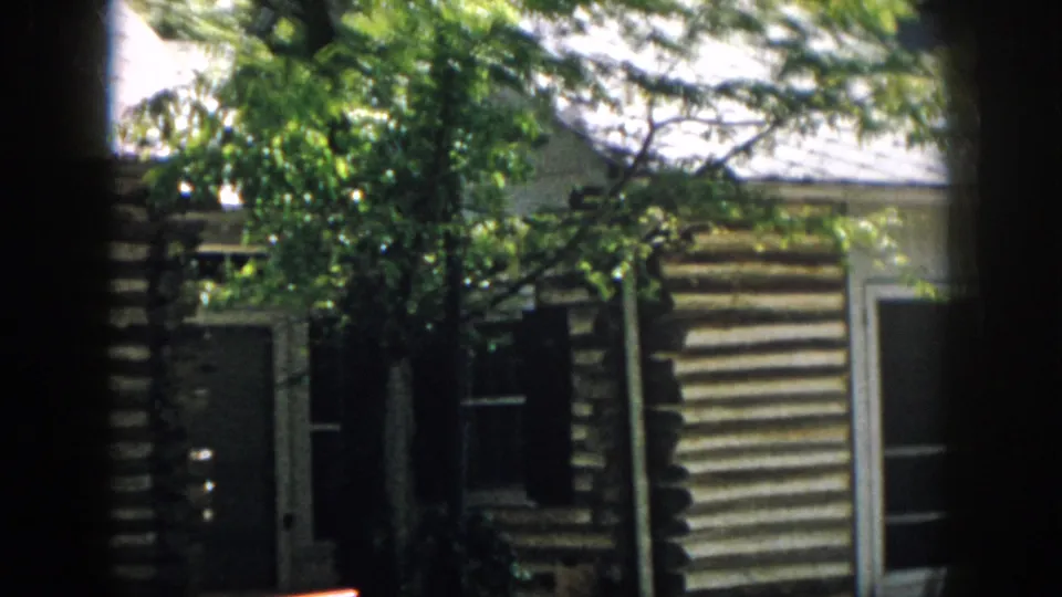 1958 Virginia home-movie frame showing a log cabin exterior partly obscured by trees and dark frame edges.