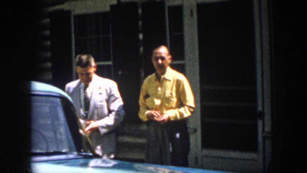 Two adult men in 1958 Virginia stand near a blue car outside a rustic cabin, one in a light suit and one in a yellow shirt.