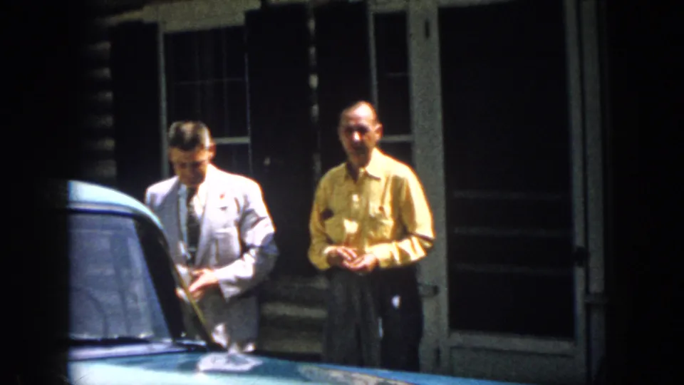 Two adult men in 1958 Virginia stand near a blue car outside a rustic cabin, one in a light suit and one in a yellow shirt.