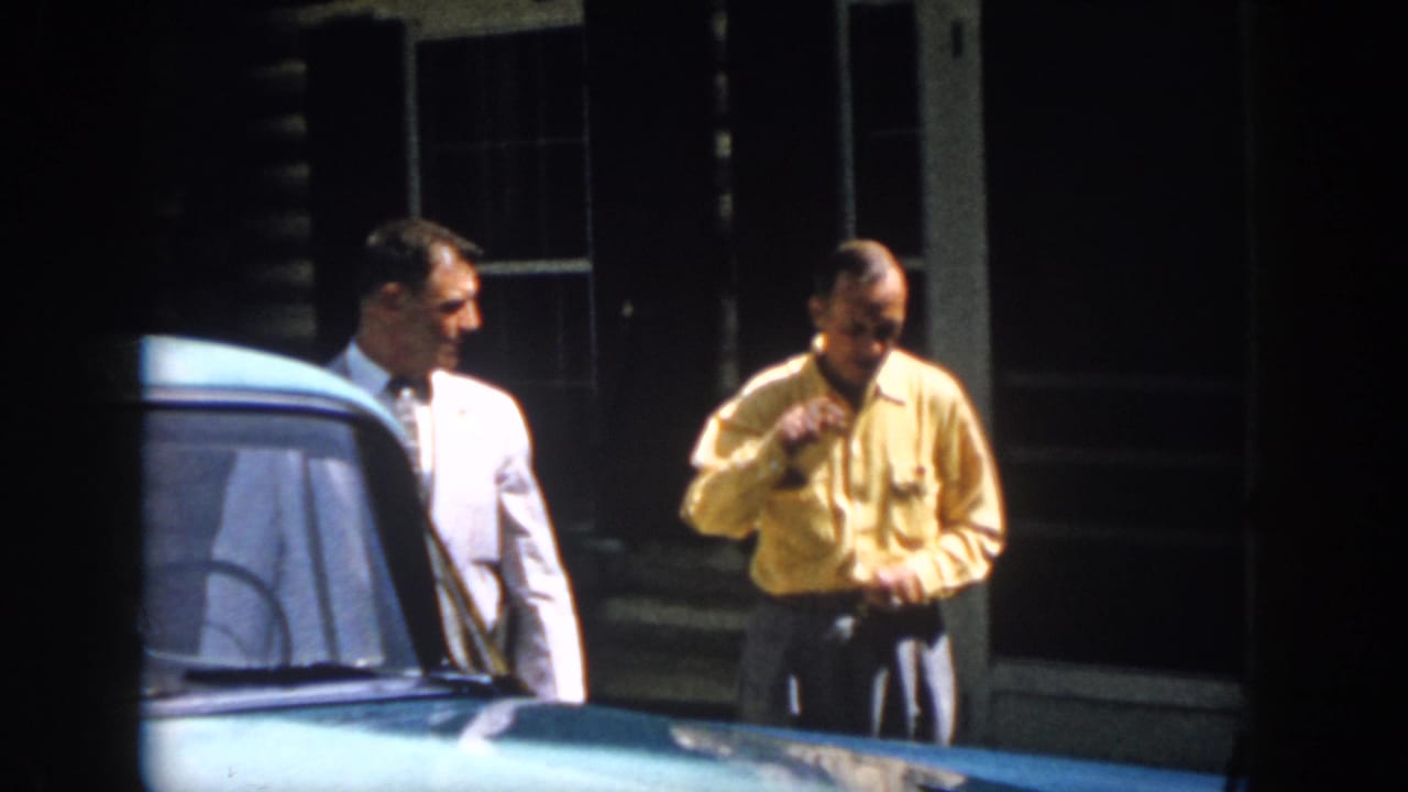Two adult men appear to converse beside a blue car outside a Virginia cabin in 1958 home-movie footage.