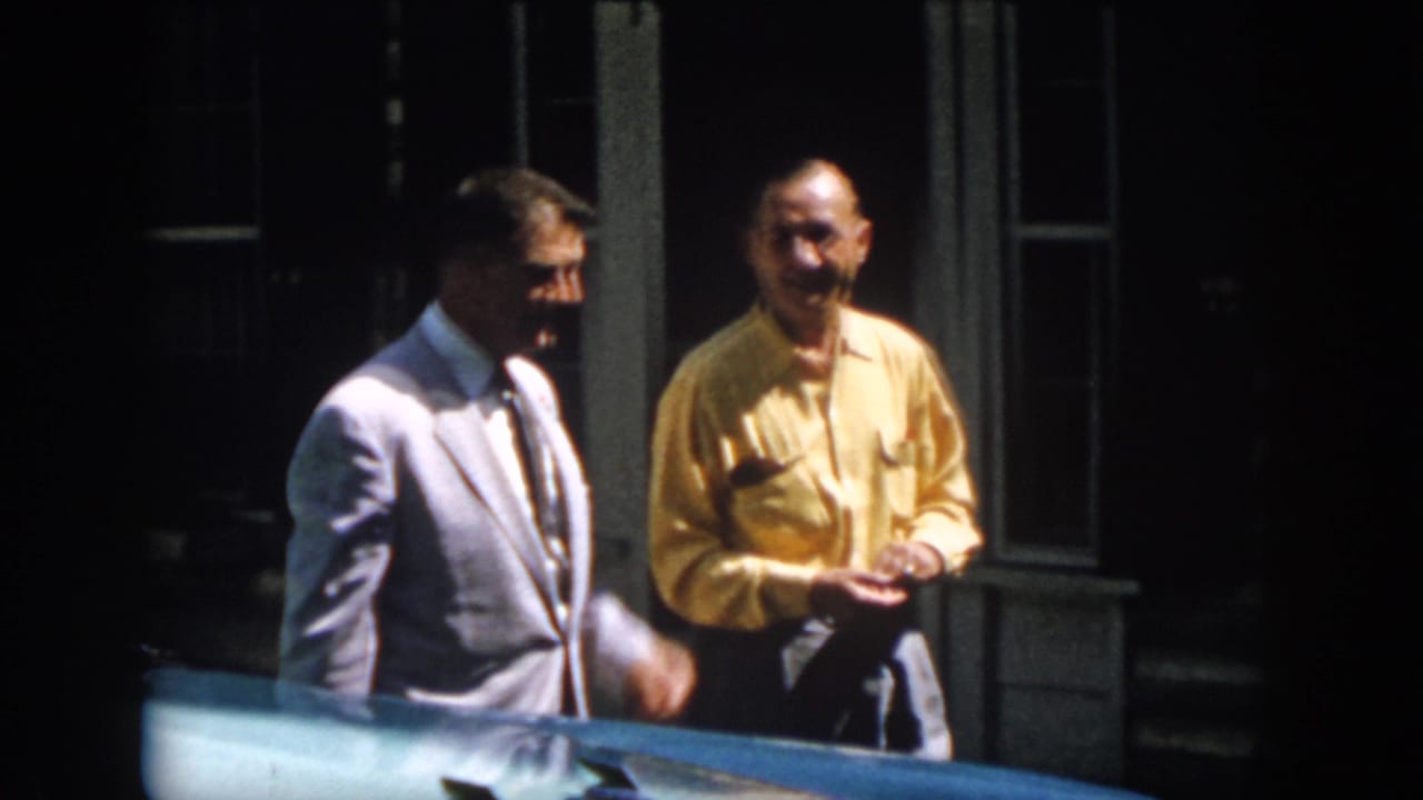 Two men smile beside a blue car outside the cabin, with shuttered windows and doorway behind them in 1958 Virginia footage.