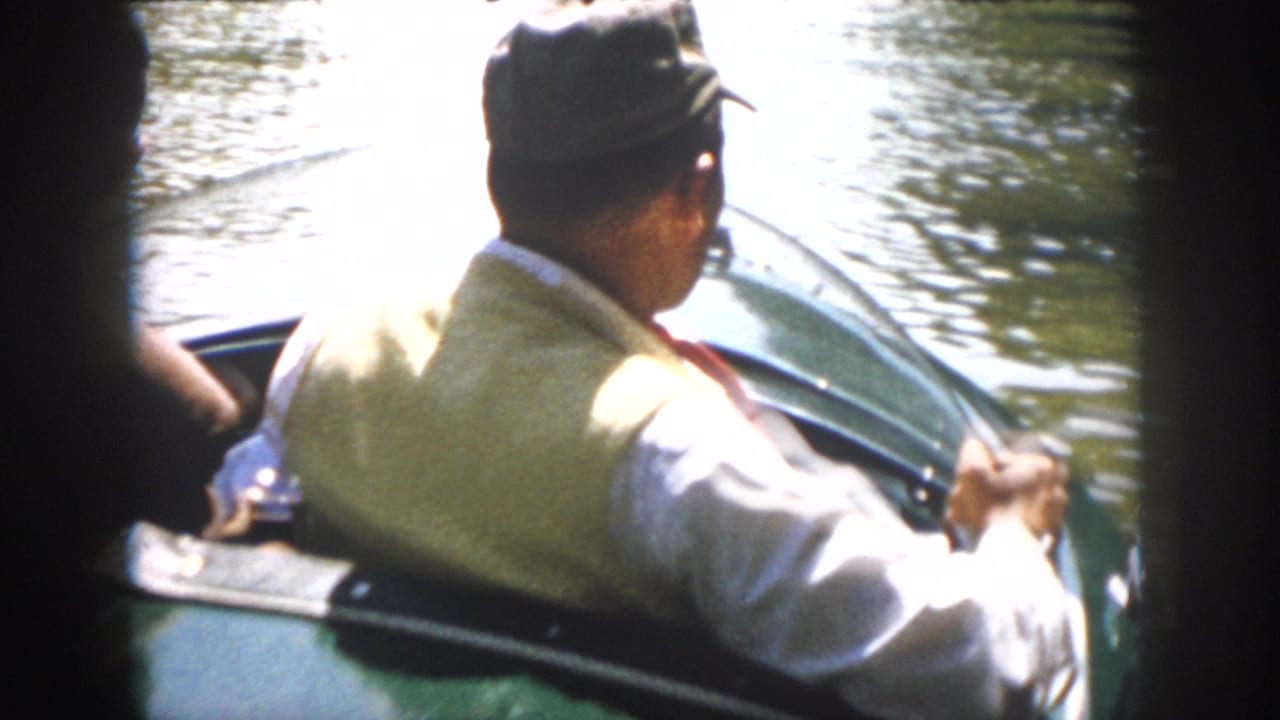 In supported 1958 Virginia home-movie footage, a man in a cap and light vest sits in a green motorboat as water reflects around him.