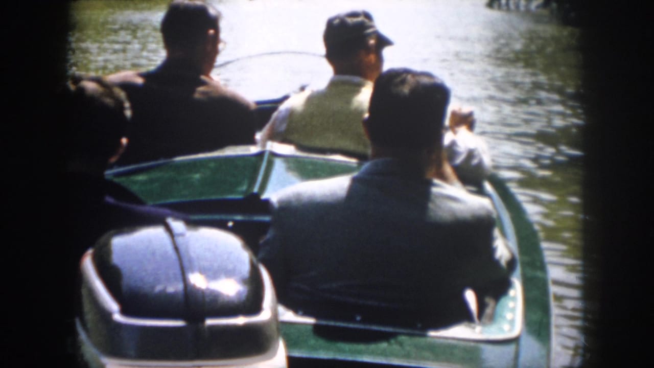 Several adult men in mid-century dress sit in a green motorboat, with the outboard motor and river visible behind them.