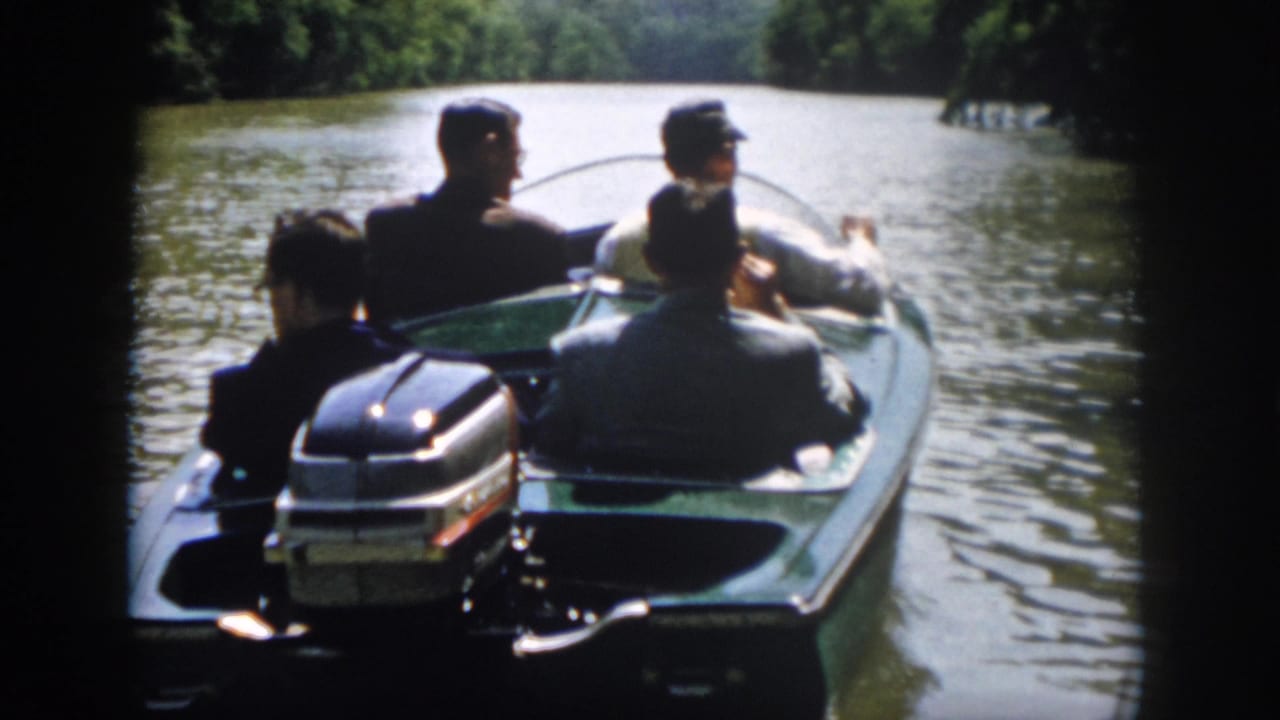 A green motorboat with several adult men aboard travels along a wooded river in supported Virginia 1958 home-movie footage.
