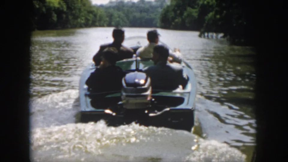 1958 Virginia 8mm frame showing four adult men in an open motorboat moving away along a tree-lined river.