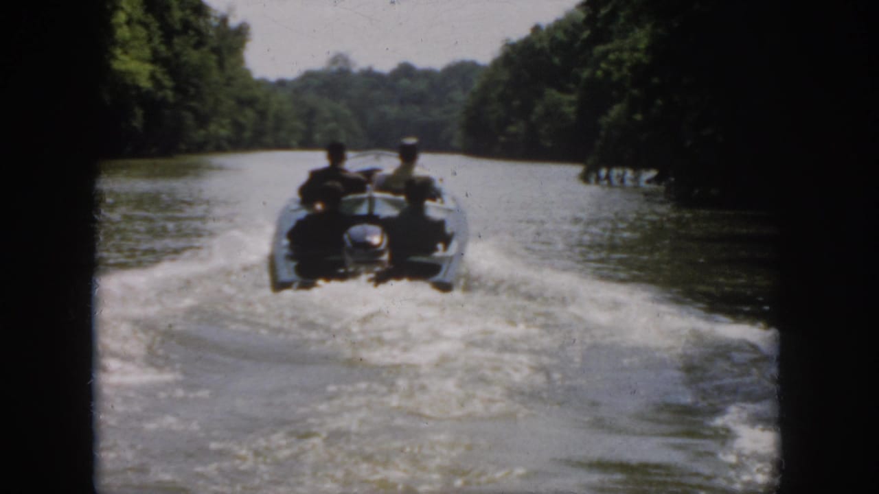 A motorboat with four men moves farther down the river between dense tree-lined banks.