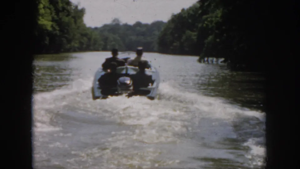 1958 Virginia home-movie frame with four men in a small boat receding along calm water and a spreading wake.