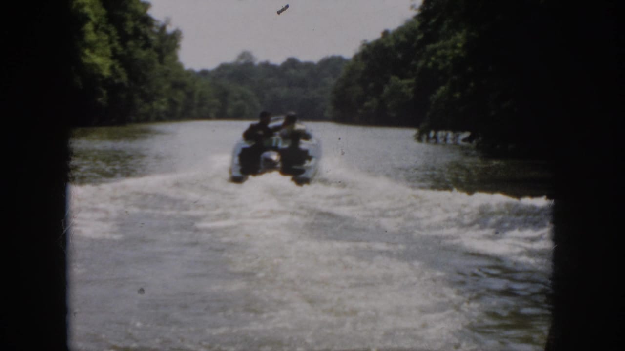 1958 Virginia 8mm frame showing a distant motorboat carrying four men along a wooded river corridor.