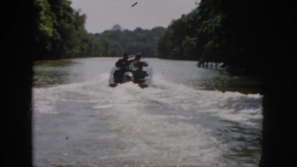1958 Virginia 8mm frame showing a distant motorboat carrying four men along a wooded river corridor.