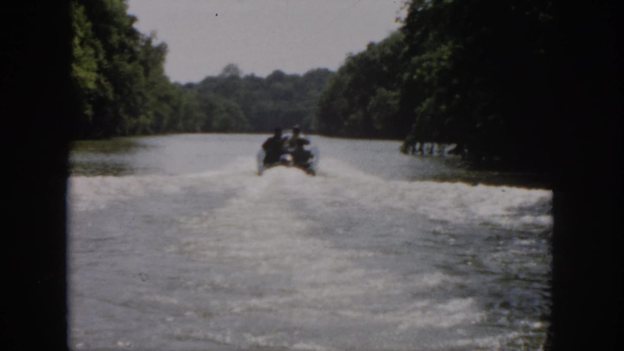 1958 Virginia home-movie frame with the motorboat far downriver, wake patterns visible on calm water between wooded banks.