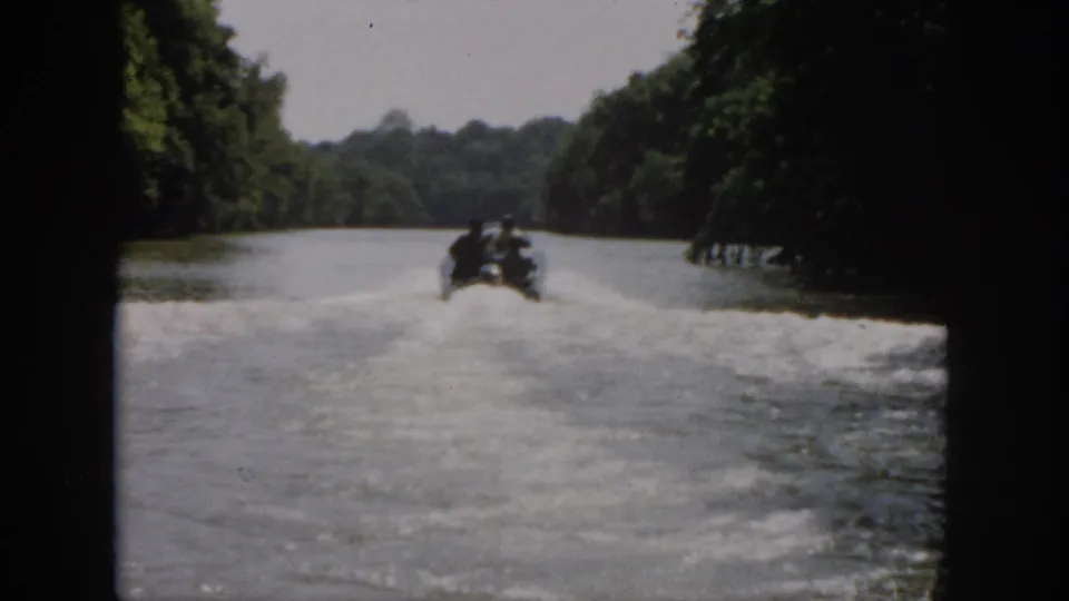 1958 Virginia home-movie frame with the motorboat far downriver, wake patterns visible on calm water between wooded banks.