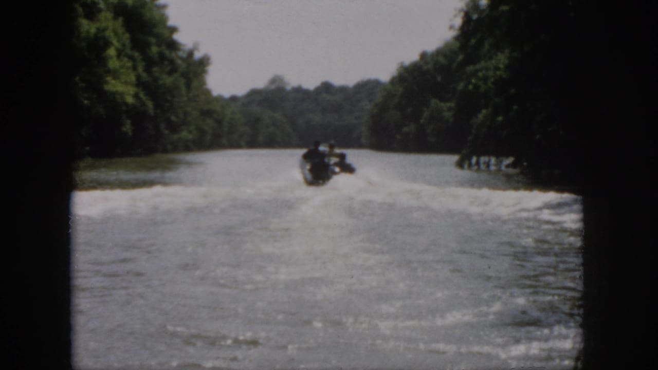 1958 Virginia 8mm frame showing a speedboat receding along a wooded river, with dark film edges and wake across the water.
