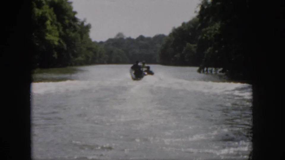 1958 Virginia 8mm frame showing a speedboat receding along a wooded river, with dark film edges and wake across the water.