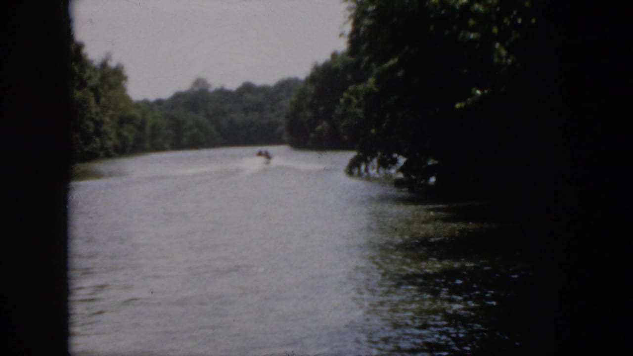 1958 Virginia home-movie frame with a small speedboat farther down a wooded river, leaving a pale wake in soft 8mm focus.