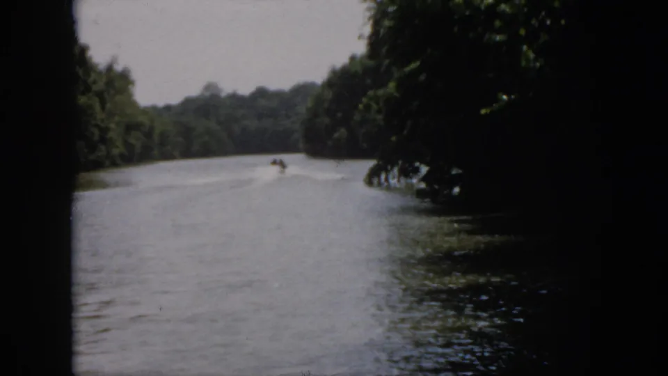 1958 Virginia home-movie frame with a small speedboat farther down a wooded river, leaving a pale wake in soft 8mm focus.