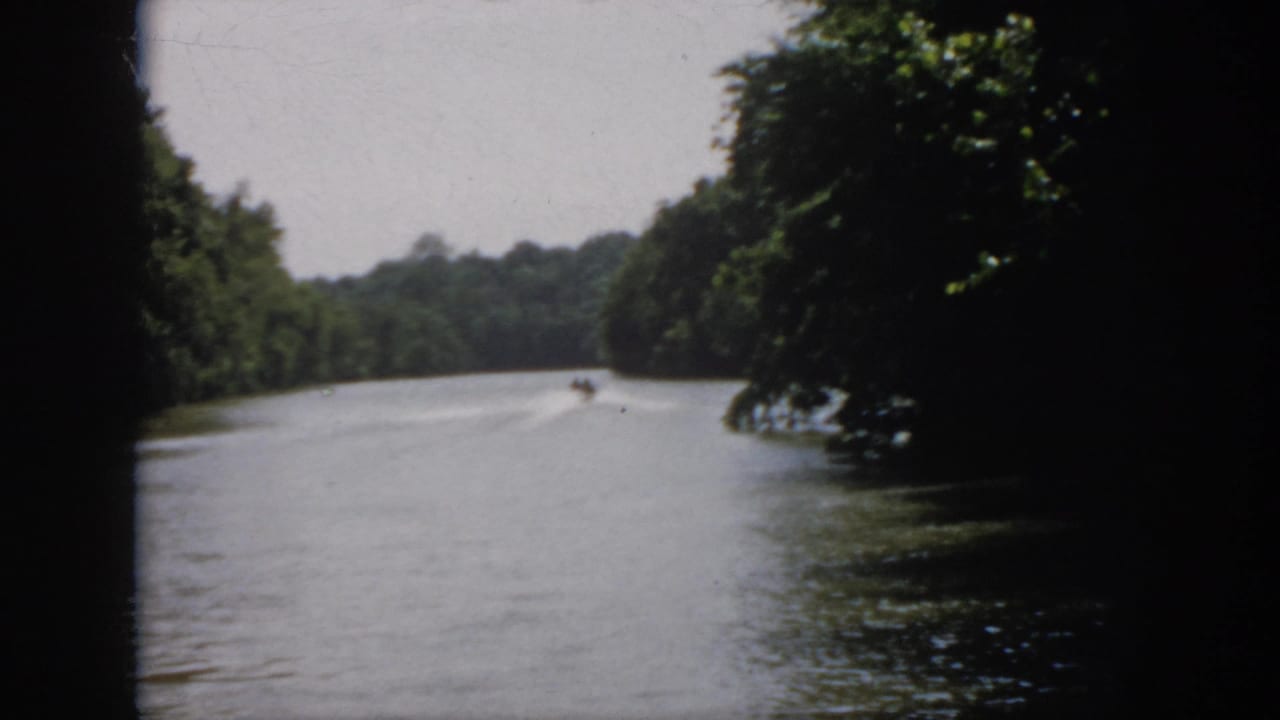 1958 Virginia 8mm frame showing the boat near the center distance on a wooded river, framed by foliage and dark film edges.