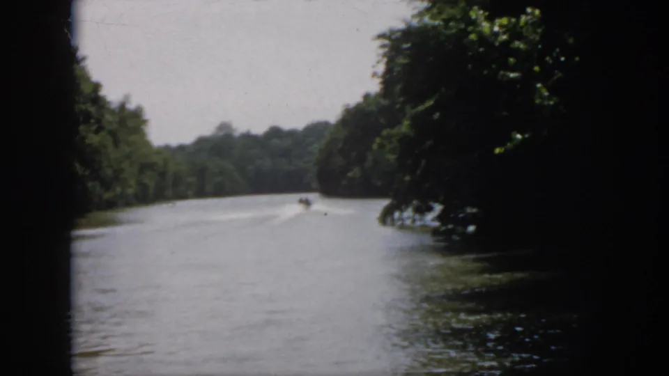 1958 Virginia 8mm frame showing the boat near the center distance on a wooded river, framed by foliage and dark film edges.