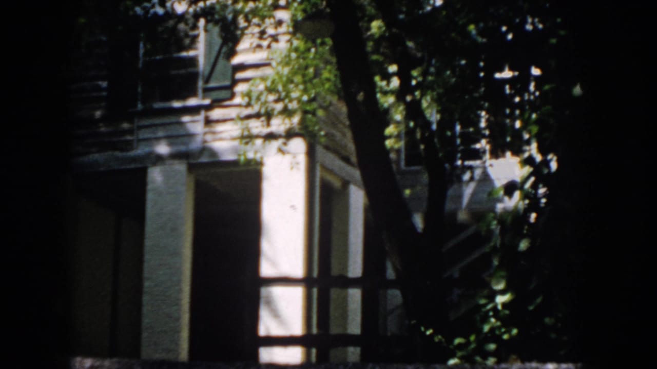 1958 Virginia home-movie frame showing a house corner and window behind riverside foliage, with deep shadow at the frame edge.