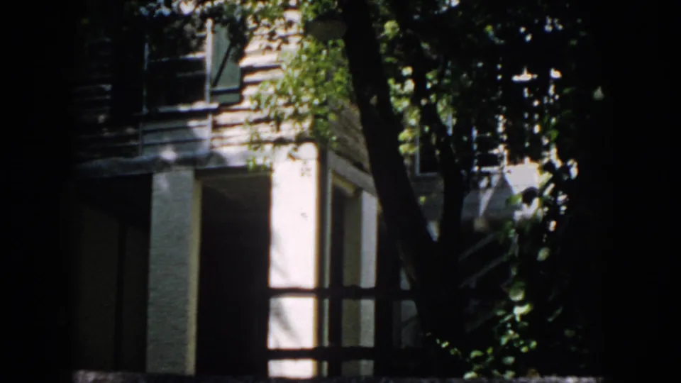 1958 Virginia home-movie frame showing a house corner and window behind riverside foliage, with deep shadow at the frame edge.