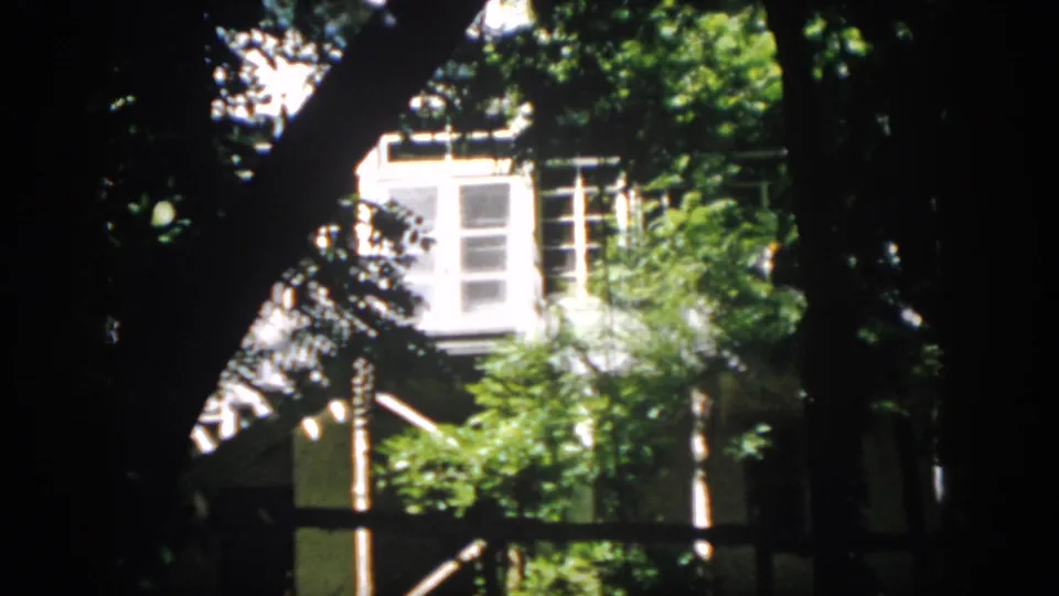 The frame shows multi-pane windows, bright siding, and leafy branches in a handheld 1958 Virginia exterior view.