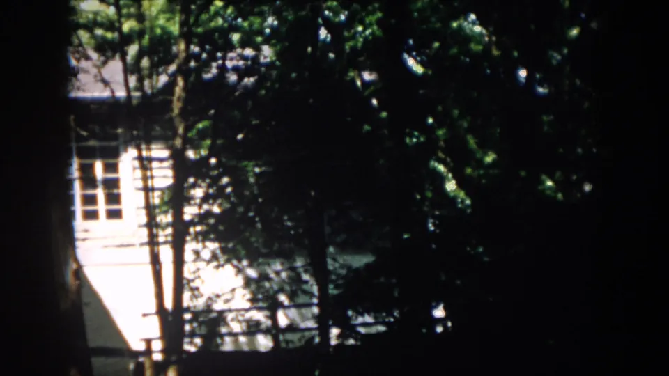 The camera view shifts lower, holding on the white residential facade behind trees in Virginia home-movie footage from 1958.