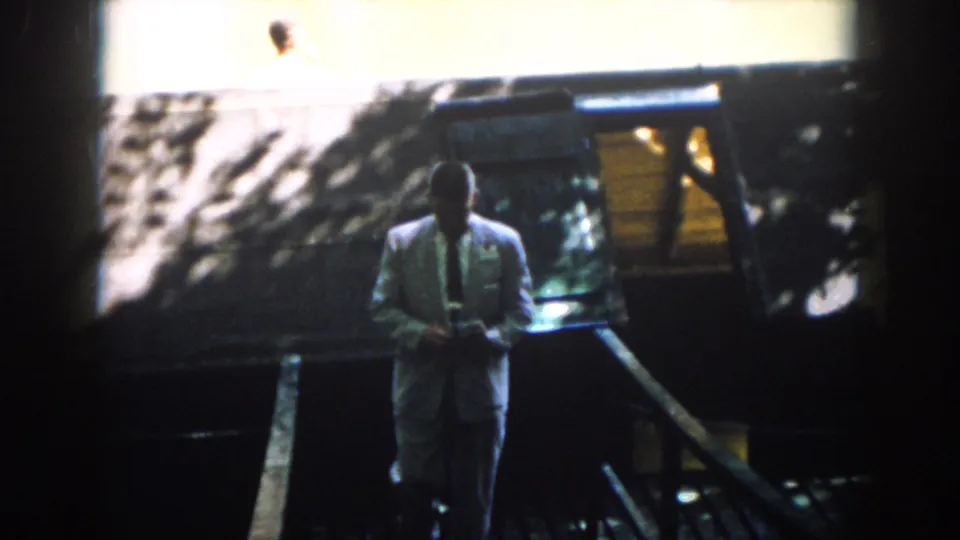 In 1958 Virginia home-movie footage, a suited man moves down a shaded wooden stair or ramp toward the camera.