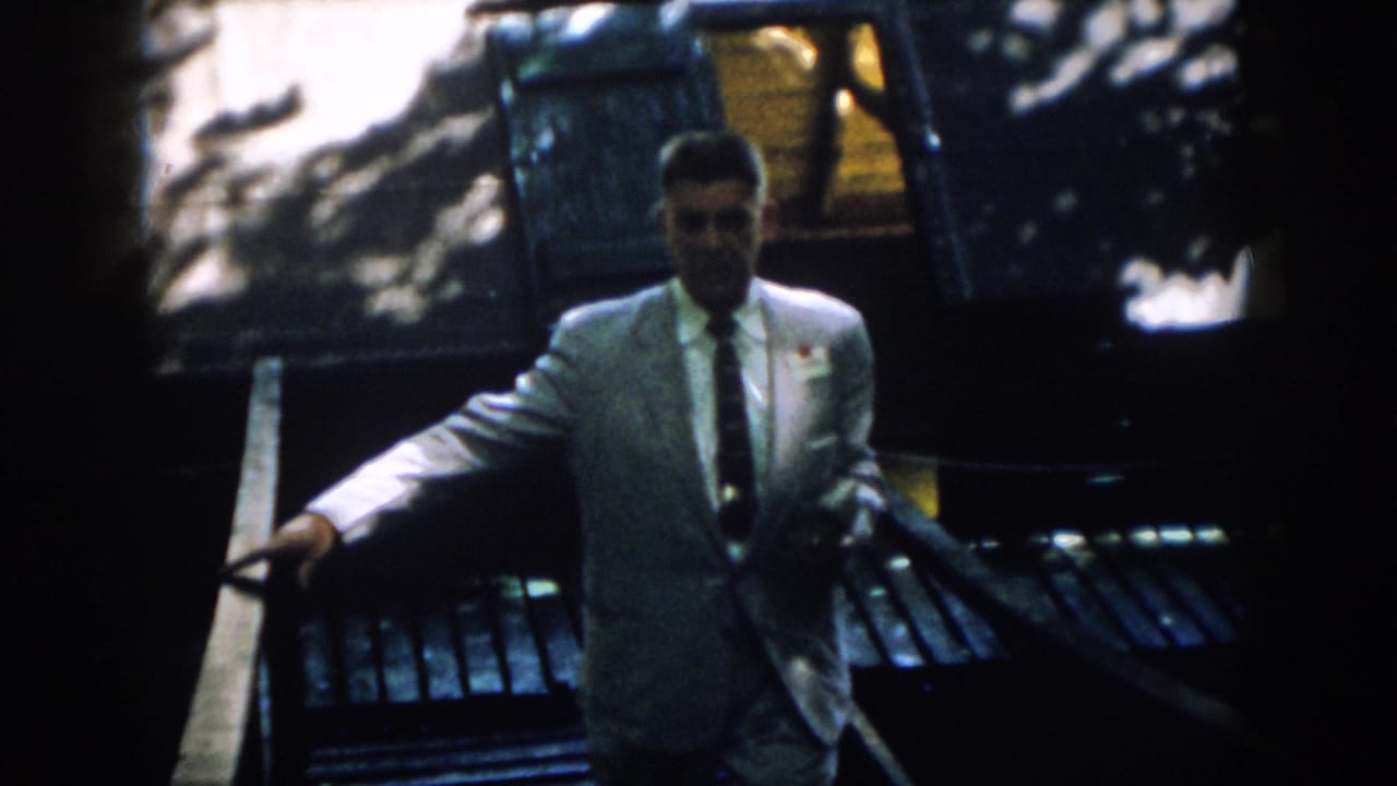 The suited man comes closer to the camera, one hand on the rail and a badge or tag visible on his jacket.