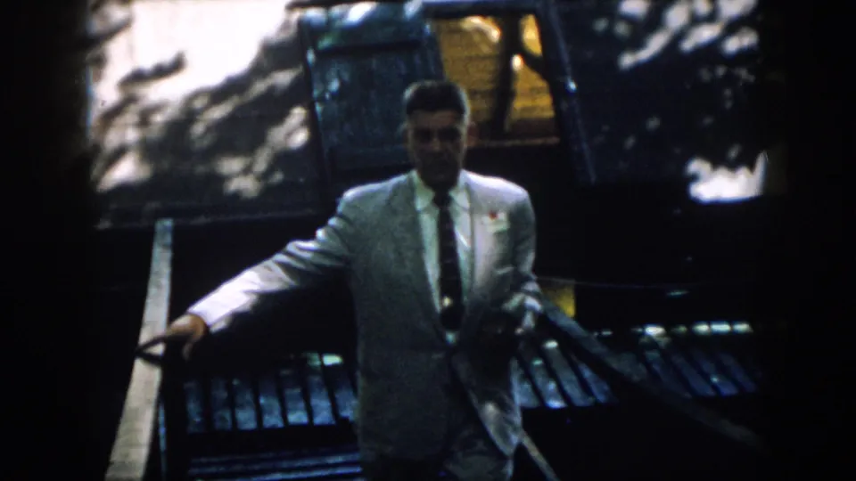 The suited man comes closer to the camera, one hand on the rail and a badge or tag visible on his jacket.