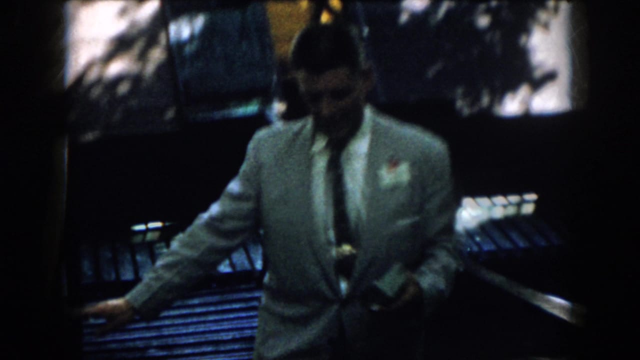 Close frame of the suited man in 1958 Virginia footage, with a possible name tag and small object near his waist.