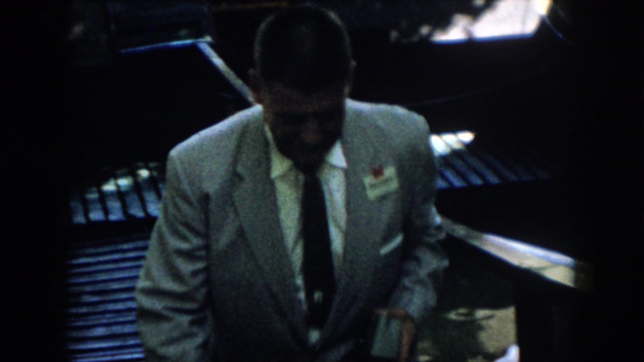 In 1958 Virginia home-movie footage, a man in a light suit and dark tie walks up a shadowed stairway with a lapel tag on his jacket.