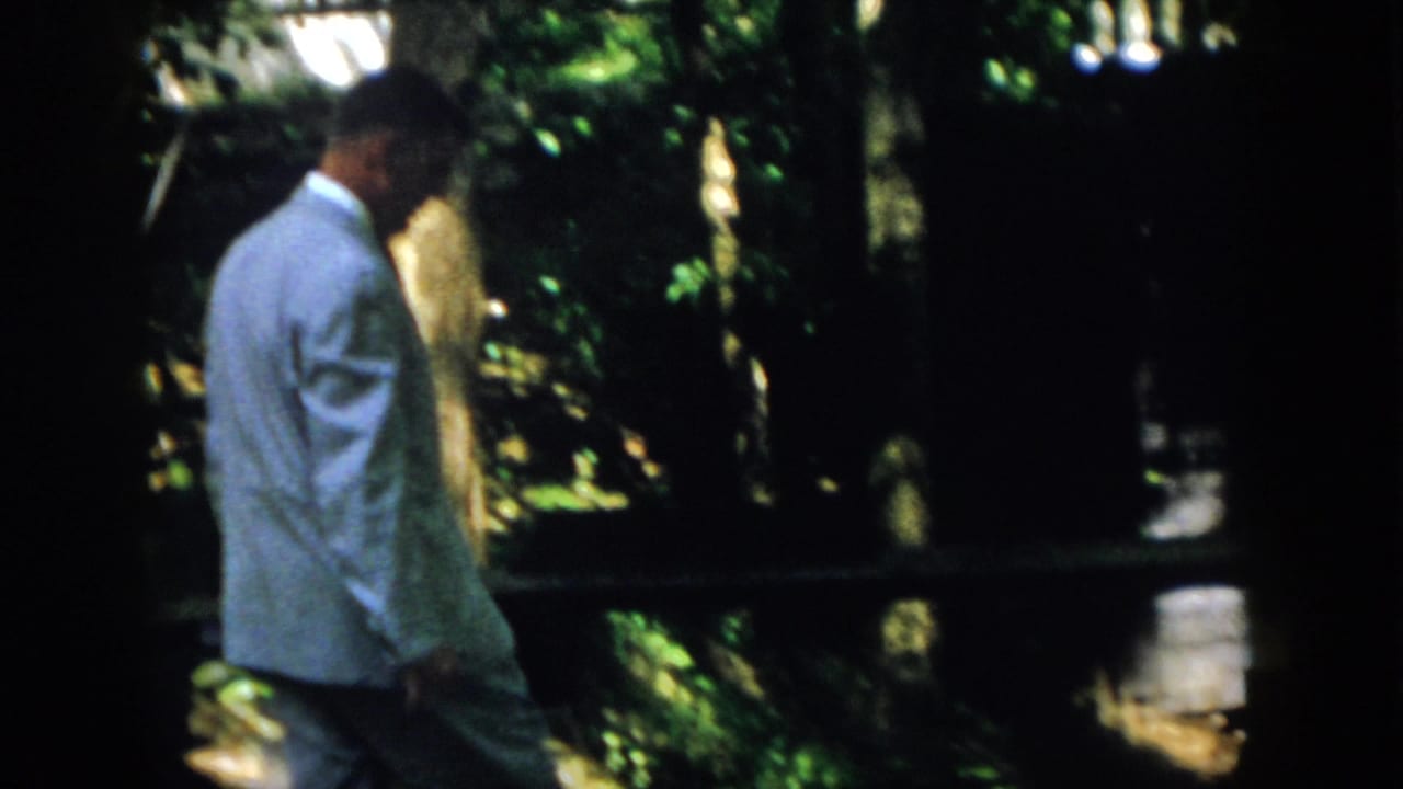 1958 Virginia home-movie frame of a suited adult man walking past shaded resort foliage and dark structural framing.