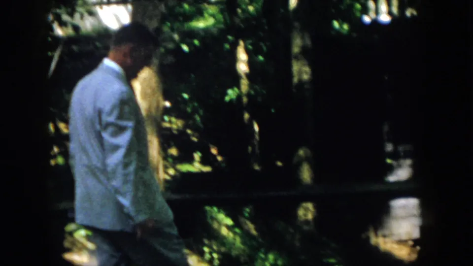1958 Virginia home-movie frame of a suited adult man walking past shaded resort foliage and dark structural framing.