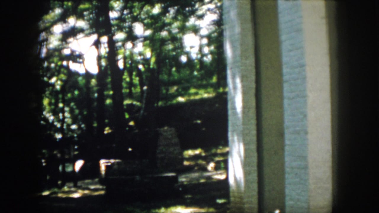 1958 Virginia 8mm frame looking from a shaded brick structure toward trees, ground shadows, and a quiet campsite area.