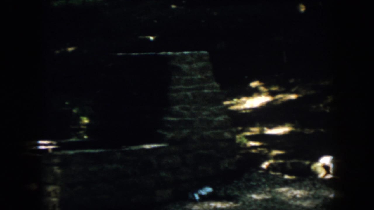 1958 Virginia 8mm frame showing a shadowed wooded campsite floor and a low stone or brick fireplace structure.