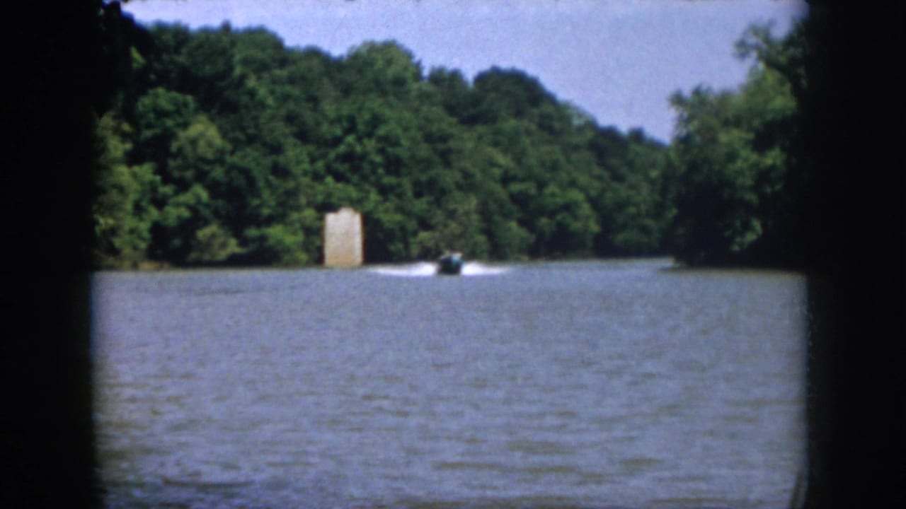 1958 Virginia home-movie frame showing a small motorboat moving across a wooded lake with a pale shoreline structure in the distance.