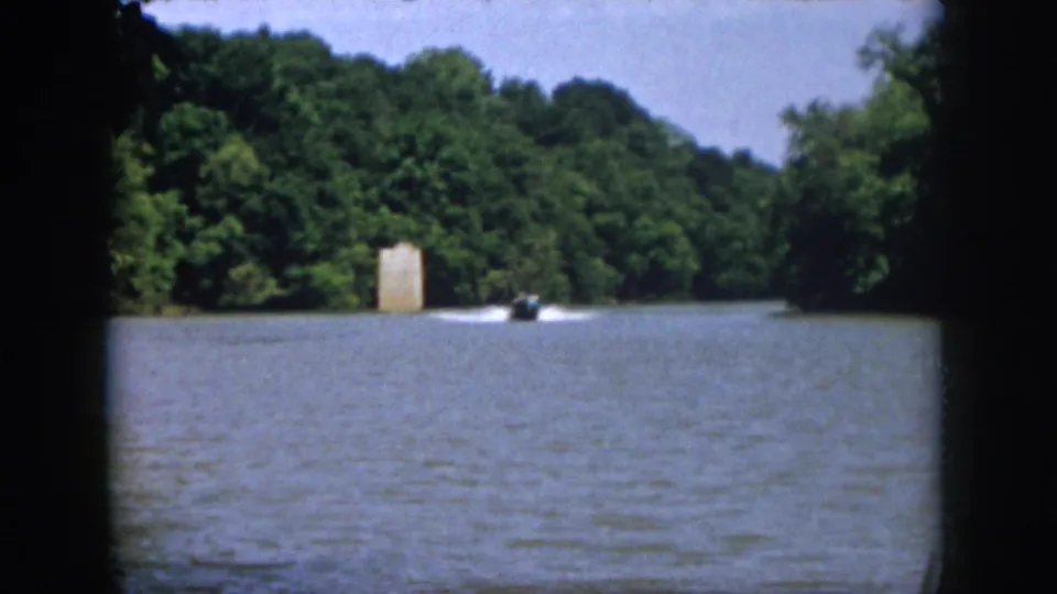 1958 Virginia home-movie frame showing a small motorboat moving across a wooded lake with a pale shoreline structure in the distance.