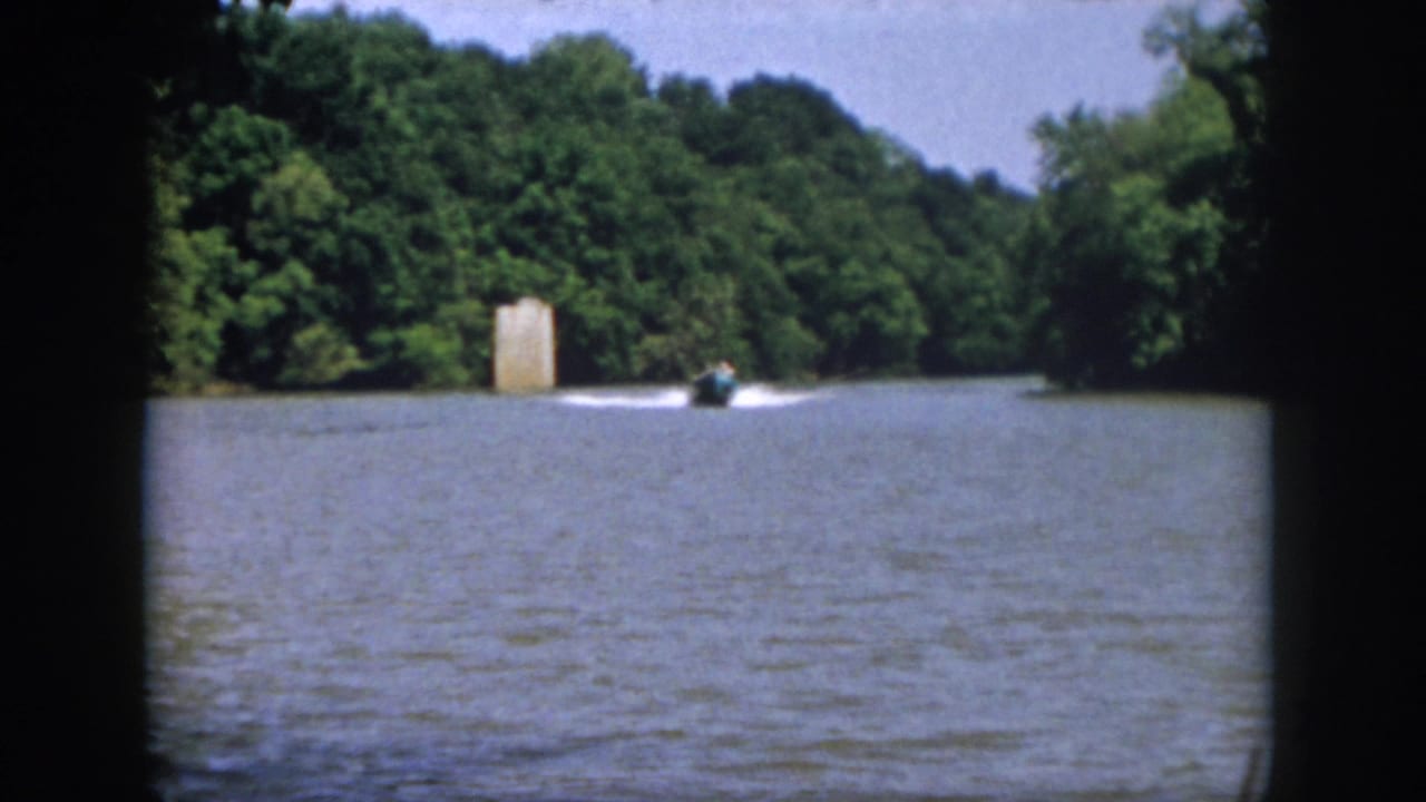 Distant 1958 Virginia lake view with a small motorboat moving past wooded shoreline and a tall pale structure near the water.