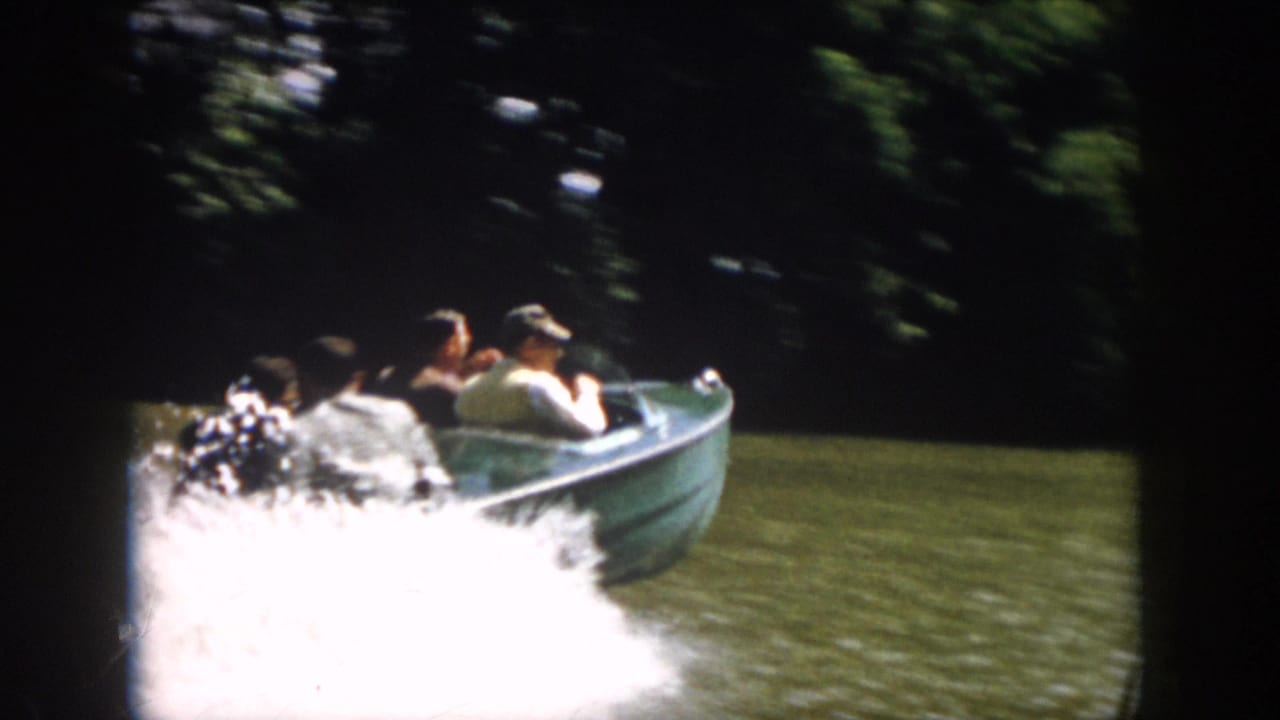 1958 Virginia 8mm frame showing a green powerboat carrying several people as it speeds along a wooded river shoreline.
