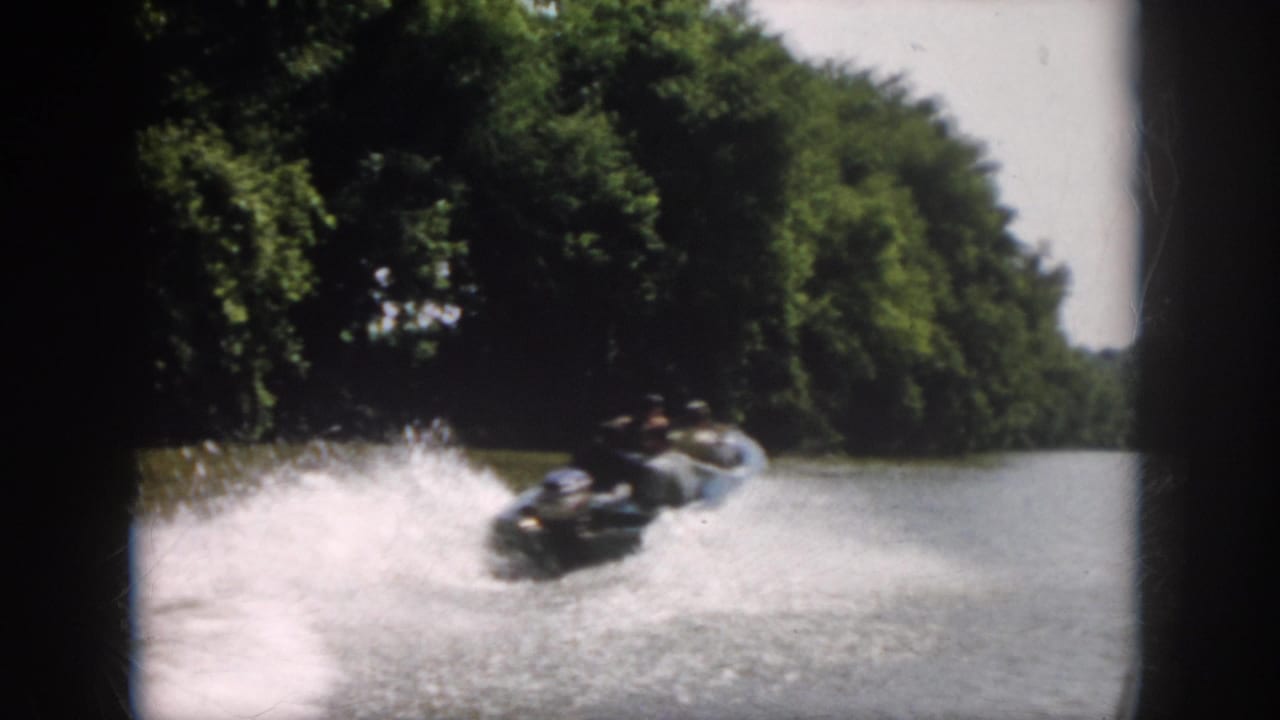 1958 Virginia home-movie frame with the powerboat receding on the river, leaving white spray against a heavily treed shoreline.
